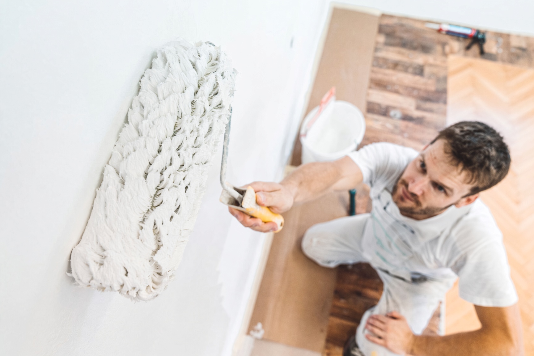 Decorator rolling white paint onto a wall, seen from above, with a paint tub on the floor beside him.